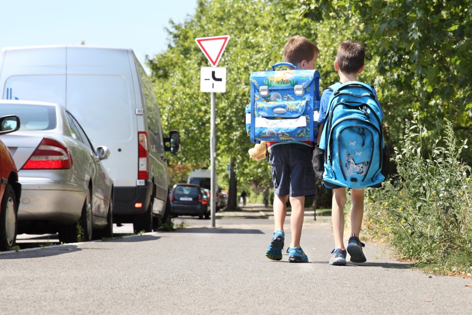 Zwei Kinder gehen einen Fußweg entlanglinks daneben steht eine Reihe geparkter Autos, im Hintergrund steht ein Vorfahrtszeichen.