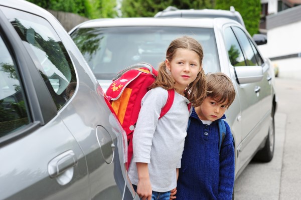 Zwei Kinder schauen vorsichtig zwischen geparkten Autos auf die Straße. 