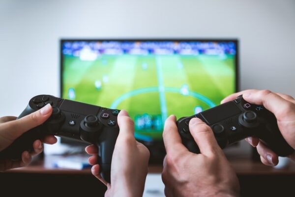Vier Hände halten zwei Controller vor einem Fernseher auf dem ein Fußballgame läuft