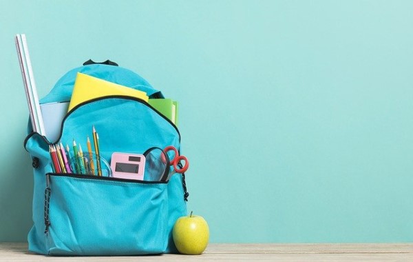 Ein Rucksack voller Schulmaterialien steht neben einem Apfel vor einer Wand.