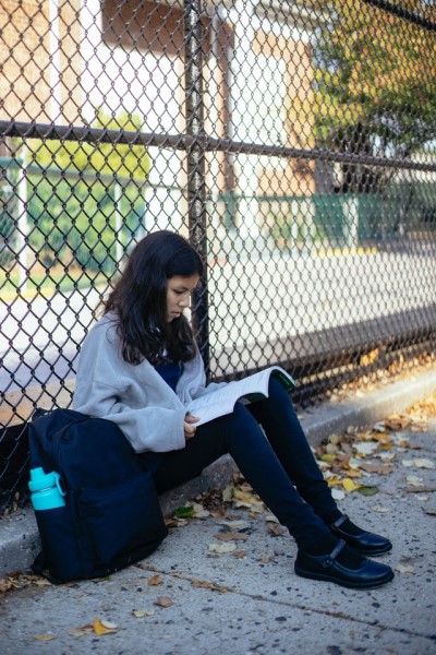 Ein Mädchen sitzt mit Schultasche am Boden vor einem Zaun und liest ein Buch. 
