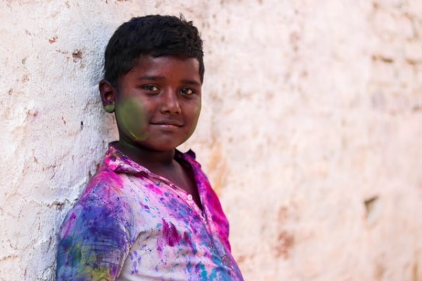 Ein Junge mit Farbflecken im Gesicht und auf der Kleidung steht lächelnd vor einer Wand.