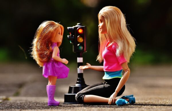 Eine erwachsene und eine kindliche Barbie Puppe schauen sich an. Zwischen ihnen steht eine kleine Spielzeug-Ampel. 