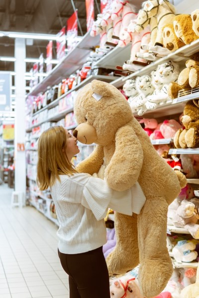 Mädchen steht vor einem Spielzeugregal und hält großen Teddy im Arm.