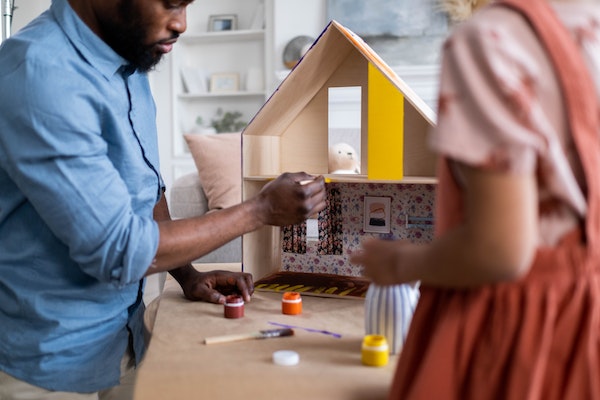 Ein Vater malt mit seinem Kind ein selbstgebautes Puppenhaus farbig an. 