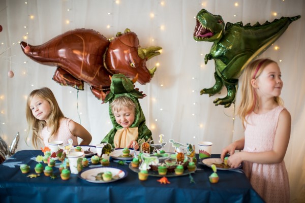 Ein Kindergeburtstagstisch mit Dino-Deko und drei Kinder, die um einen Tisch mit Muffins stehen.