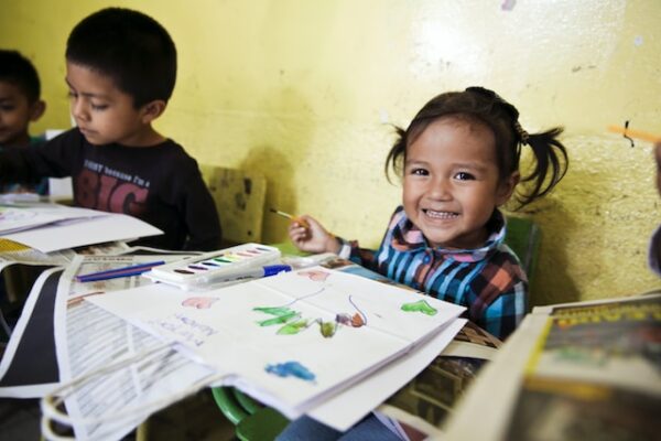 Ein Vorschulkind lächelt der Kamera zu, während es auf einem Blatt Papier malt. 