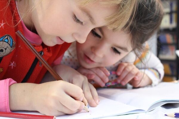 Ein Kind schreibt in ein Heft, während ein jüngeres Kind interessiert zuschaut. 