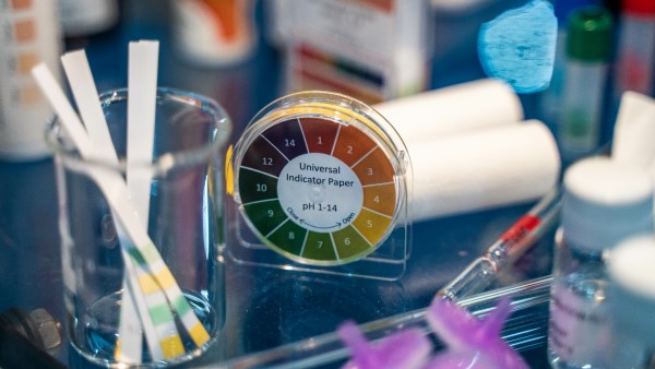 Benutzte pH-Indikatorpapierstreifen stehen in einem Glasbecher auf einem Tisch voller Labor-Equipment. Daneben liegt eine frische Rolle Indikatorpapier inklusive Farbanzeige für die jeweiligen pH-Werte. 