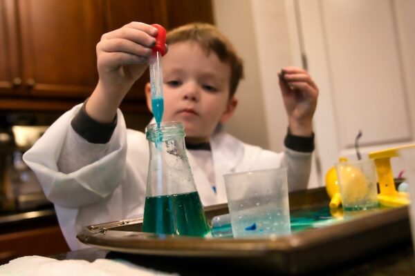 Ein Kind in weißem Laborkittel hält eine Pipette, mit dem es eine farbige Flüssigkeit aus einem Becher aufnimmt. 