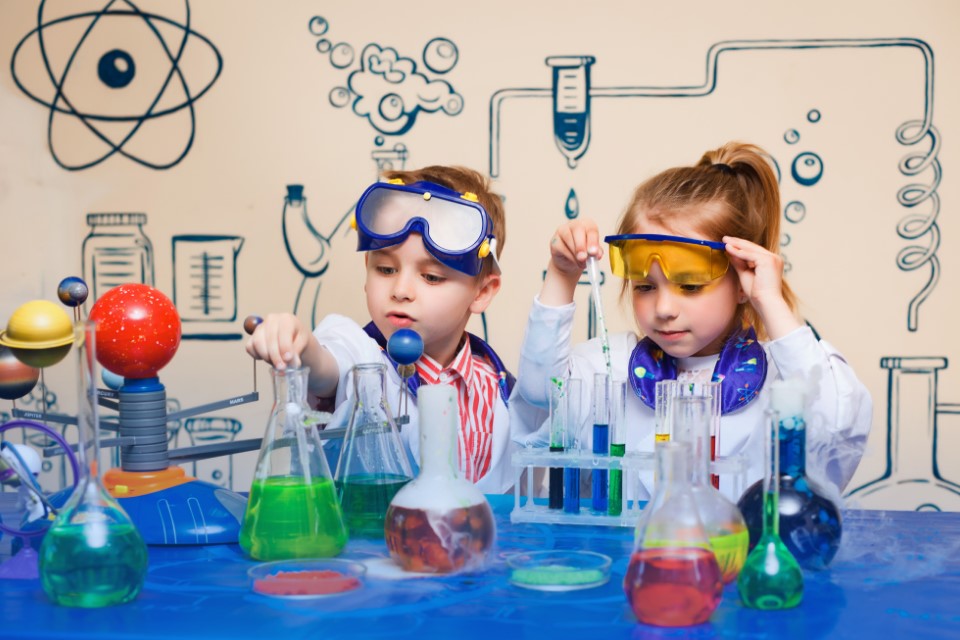 Zwei Kinder in Schutzkleidung sitzen vor einem Tisch und experimentieren mit chemischen Flüssigkeiten.