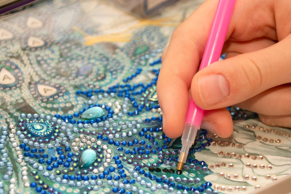 Eine Nahaufnahme von einem blauen Diamond Painting, auf das gerade eine Person mit einem Stift Perlen anbringt.